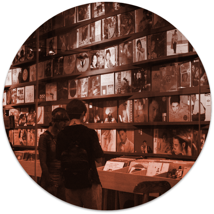 Two people browsing through a record store, there is a large wall of LP records of various artists, the image has a faint orange filter.