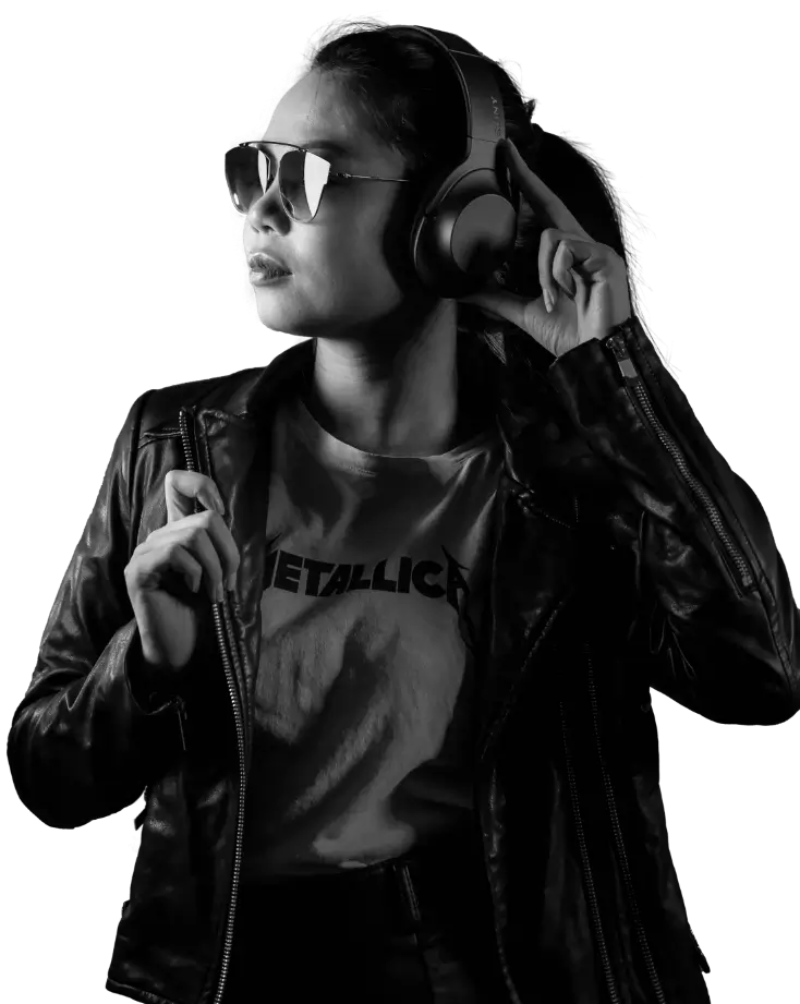 In black and white, a black haired woman in a leather jacket, sunglasses and wearing large headphones, facing her left. She's holding one of the ears of her headphones with one hand and the lapel of her jacket with the other. Her shirt has the logo of metal band "Metallica".
